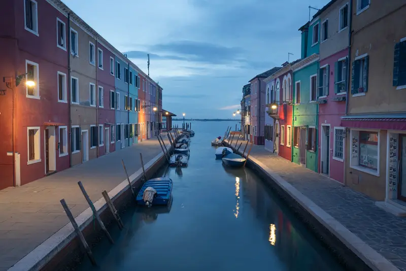 Colourful canal lined with fishing boats and pastel houses at blue hour in Burano, Venice.