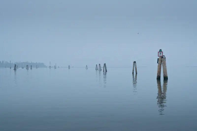 Navigation markers emerging from calm waters in the Venetian Lagoon near Burano on a misty morning.