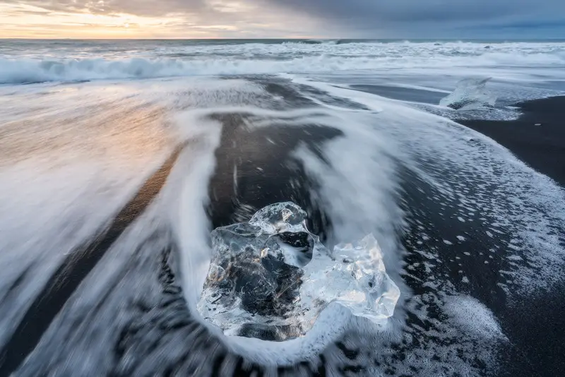 Flowing waves surrounding a crystal-clear ice block on Diamond Beach, Iceland.
