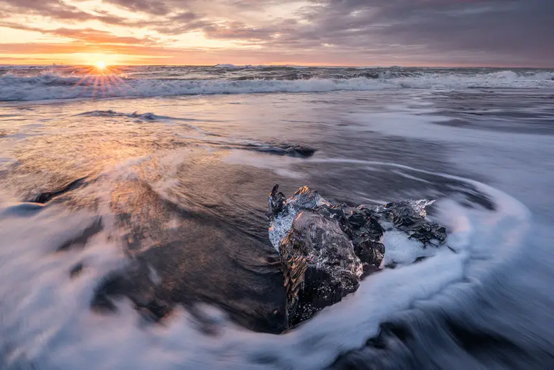 Dark ice block surrounded by flowing waves at sunrise on Diamond Beach, Iceland.
