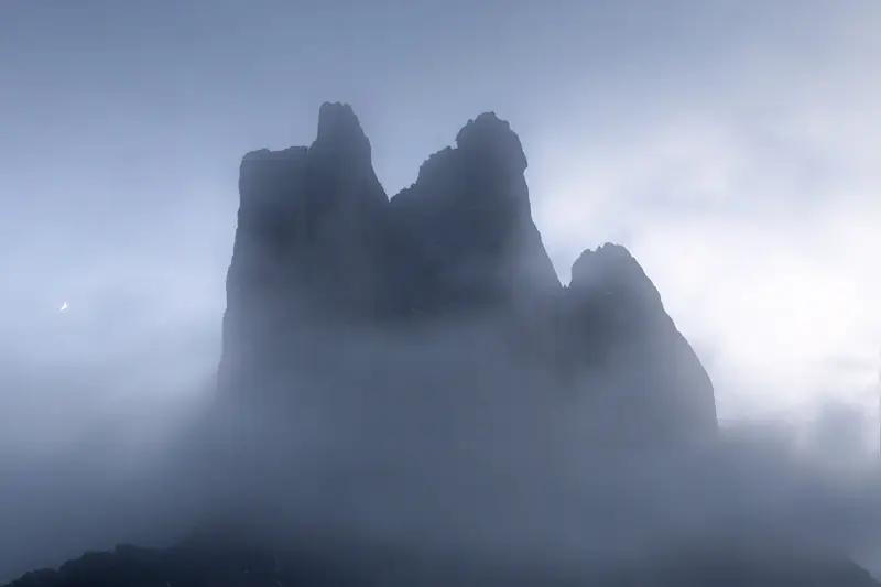Tre Cime di Lavaredo emerging through dense fog with a crescent moon in the Dolomites