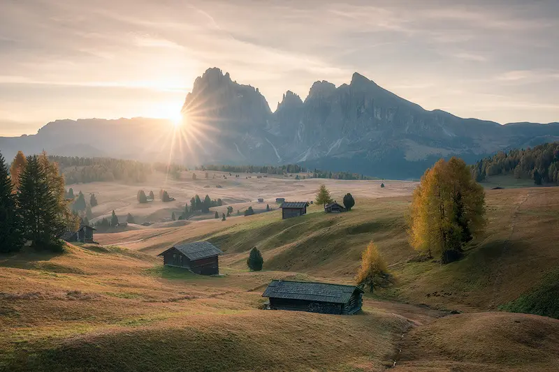 Autumn sunrise over Alpe di Siusi in the Italian Dolomites, with golden sunlight illuminating alpine meadows, wooden huts and the Sciliar mountains.