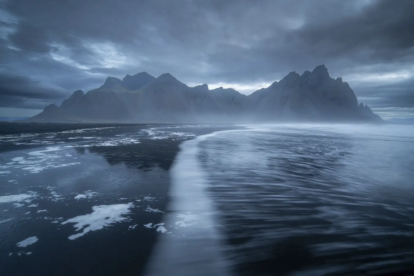 Vestrahorn mountain reflected on the wet black sand beach of Stokksnes, Iceland, with curved ocean waves and dramatic blue hour clouds