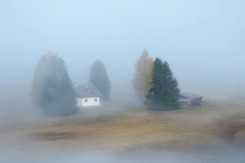 Traditional alpine cabins and autumn trees surrounded by dense fog on Alpe di Siusi in the Italian Dolomites