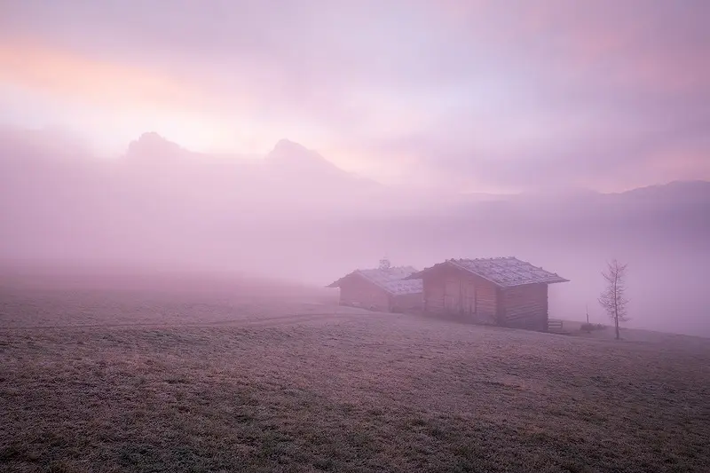 Traditional wooden alpine huts surrounded by pink morning mist at Alpe di Siusi, with faint mountain silhouettes in the Italian Dolomites