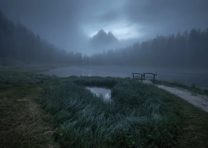 Lago d'Antorno in the Dolomites surrounded by blue mist, with a small wooden bridge and distant mountain peaks emerging through the fog