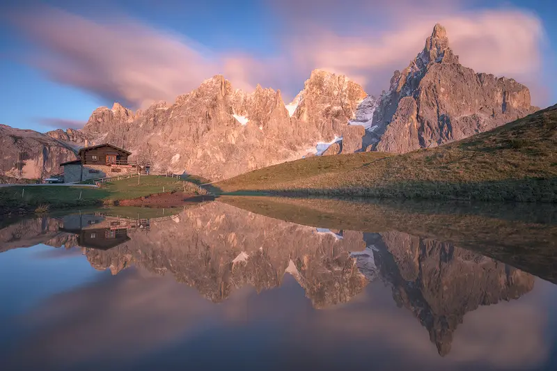 Baita Segantini at Passo Rolle reflected in a mountain pond at sunset, with the Pale di San Martino peaks in the Italian Dolomites
