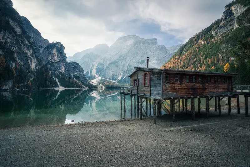 Wooden boathouse at Lago di Braies surrounded by autumn forests and dramatic Dolomites peaks reflected in the lake.