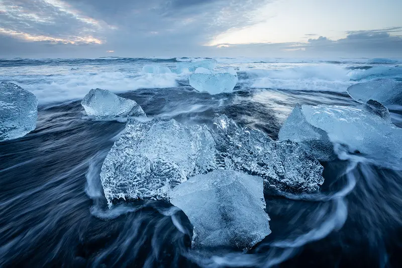 Crystal-clear glacier ice surrounded by flowing Atlantic waves on Diamond Beach, Iceland, beneath a soft blue morning sky.
