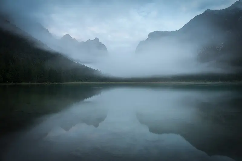 Lago di Dobbiaco in the Dolomites with low clouds drifting between mountain peaks and soft reflections on the still water