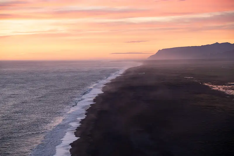 Sunset view from Dyrhólaey in South Iceland, with pastel pink skies, a sweeping black sand beach and distant mountains fading into mist.