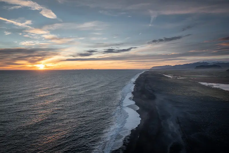 Panoramic view from Dyrhólaey over Iceland's black sand coastline at sunset, with Atlantic waves and distant mountains.