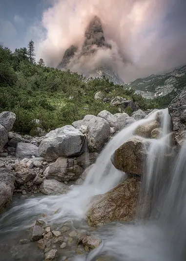 Hidden alpine waterfall flowing over rocks beneath a dramatic mountain peak surrounded by clouds in the Italian Dolomites
