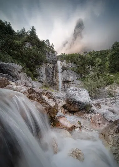 Hidden waterfall flowing over rocky terrain beneath mist-covered mountain peaks in the Italian Dolomites