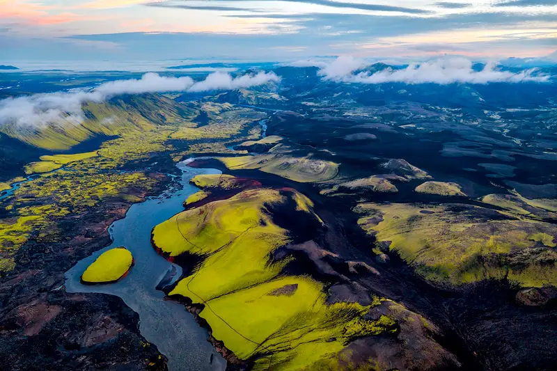 Aerial view of moss-covered volcanic hills and a winding river beneath low clouds in the Icelandic Highlands.