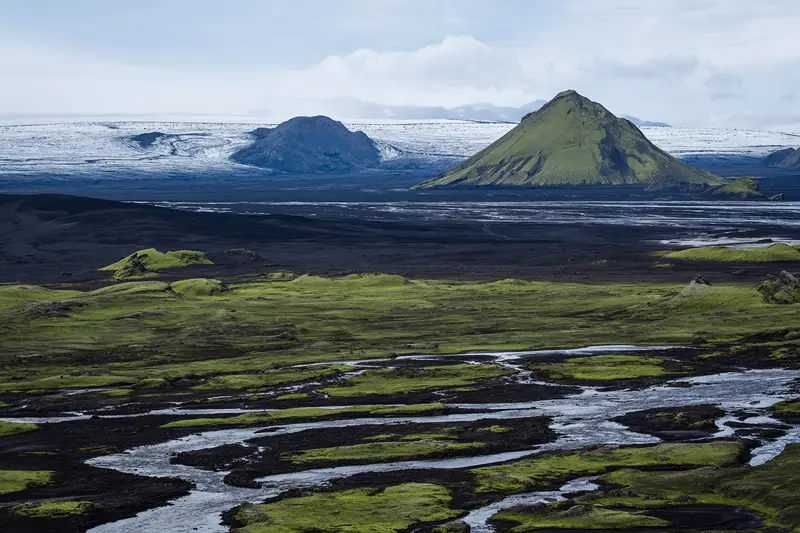 Green volcanic cone rising above black lava plains, with braided streams and a glacier in the Icelandic Highlands.