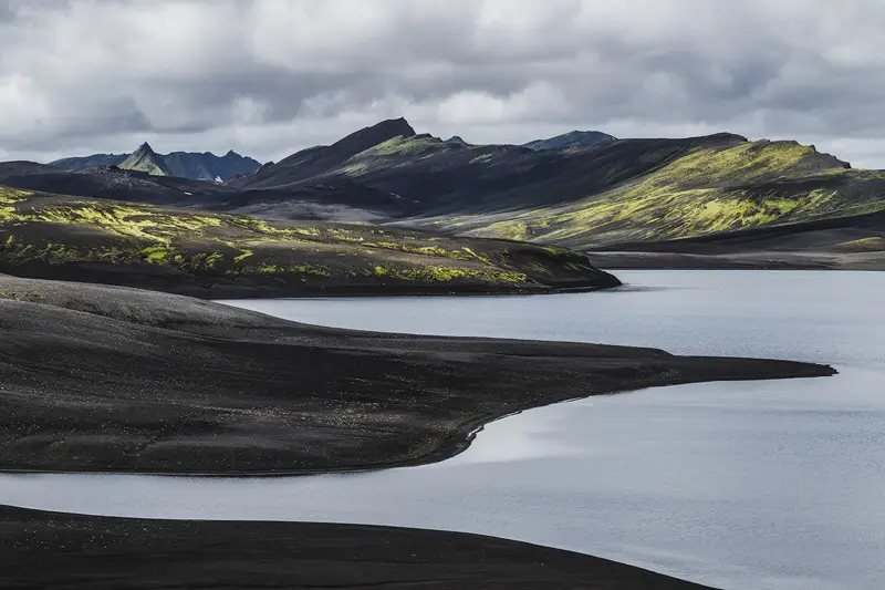 Black volcanic sand shoreline curving around a peaceful lake, with moss-covered mountains and storm clouds in the Icelandic Highlands.