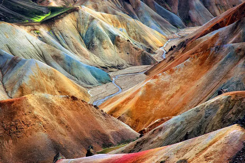 Colorful rhyolite mountain slopes in Landmannalaugar, Iceland, with orange, blue and ochre textures surrounding a narrow stream.
