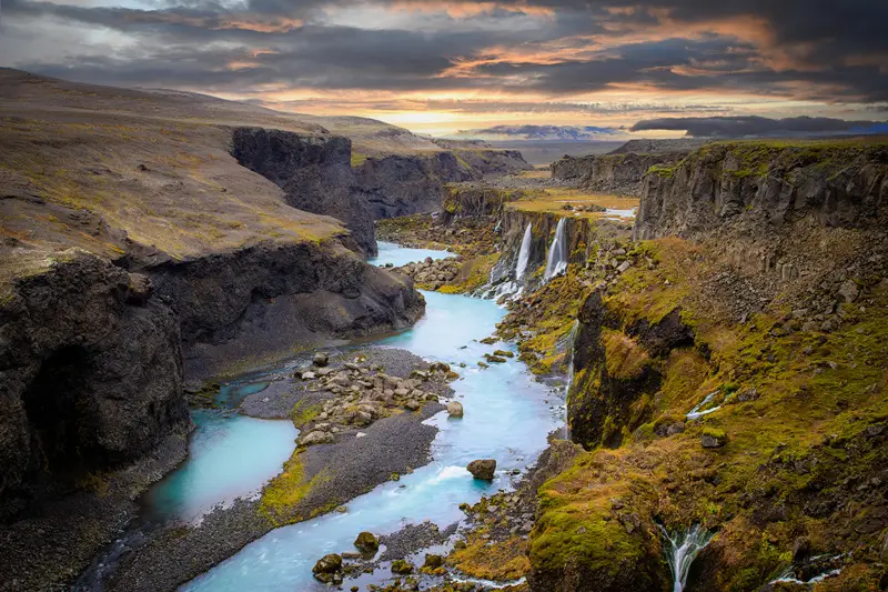 Turquoise glacial river flowing through Sigöldugljúfur canyon, with waterfalls cascading down moss-covered cliffs beneath a dramatic sky in Iceland.