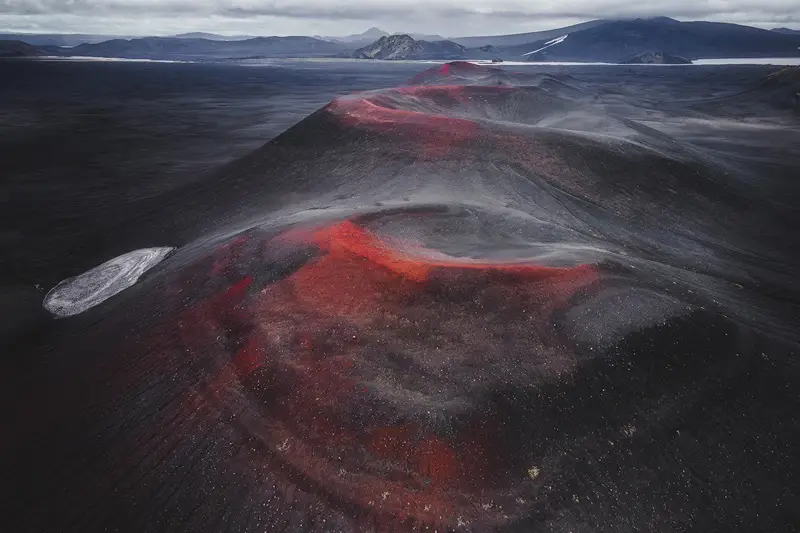 Aerial view of a chain of dark volcanic craters with red mineral tones stretching across the black volcanic landscape of the Icelandic Highlands.