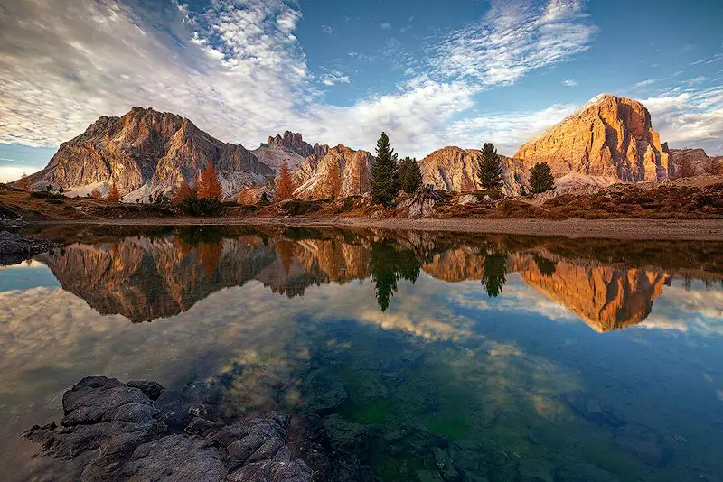 Lago di Limides in the Dolomites reflecting sunlit mountain peaks, autumn larch trees and a blue sky with scattered clouds