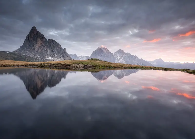 Ra Gusela and surrounding Dolomites peaks reflected in a still alpine pond at Passo Giau beneath a dramatic cloudy sky.