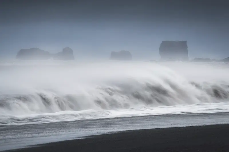 Long exposure of Atlantic waves flowing across Reynisfjara black sand beach with distant sea stacks emerging through the mist in Iceland