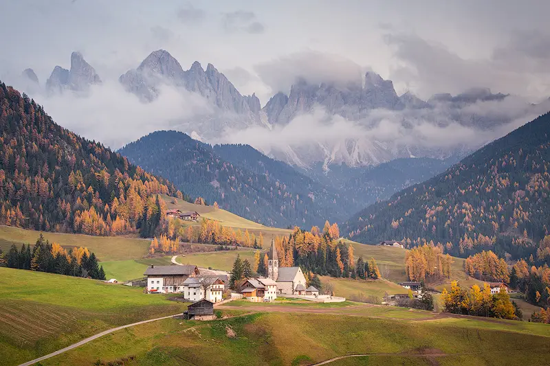 Santa Maddalena village and church in Val di Funes, surrounded by golden autumn larches beneath the cloud-covered Odle peaks in the Dolomites