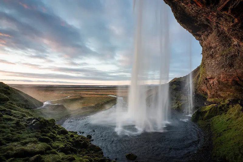 View from behind Seljalandsfoss waterfall in South Iceland, with cascading water, moss-covered cliffs and a soft evening sky over the coastal plains.