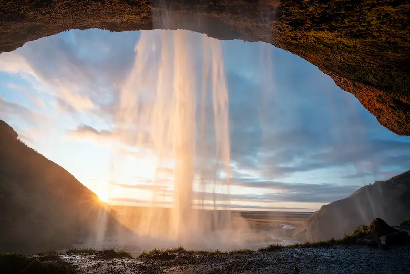 Golden sunset light shining through Seljalandsfoss waterfall in South Iceland, viewed from behind the cascading water beneath a natural rock overhang.