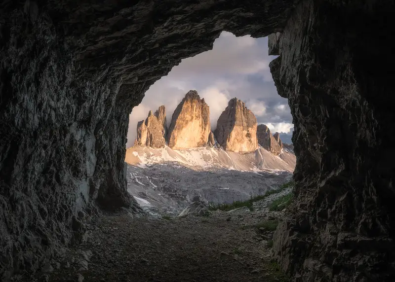 Tre Cime di Lavaredo illuminated by golden sunlight, framed by a rocky cave in the Italian Dolomites